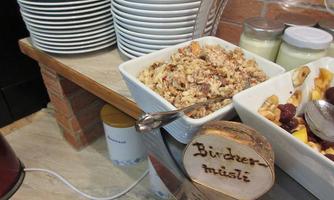 A bowl of Bircher muesli is next to stacked plates. Behind them are glasses with yogurt and a selection of fresh fruit.