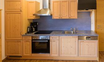 A modern kitchen with wooden cabinets and glossy surfaces. It features a stove, a sink, and a built-in TV.