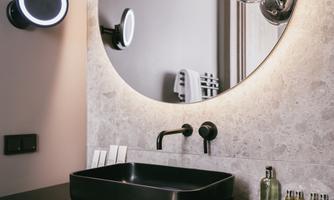 A modern bathroom with a large round mirror and an elegant black sink. The walls are in neutral tones, and there is stylish lighting.