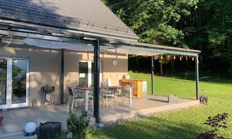 A cozy terrace with a dining table and chairs, surrounded by green grass and trees. The sun rays illuminate the space and create an inviting atmosphere.