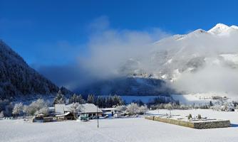 A picturesque winter landscape with snow-covered fields and mountains in the background. The sky is clear and blue, while some clouds drift over the mountains.