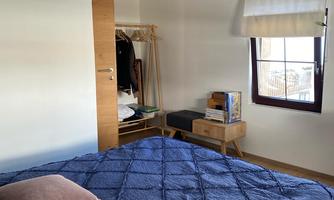 A cozy bedroom with a large bed and several pillows. There is a wardrobe and a small table next to a window.