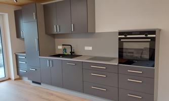 A modern kitchen with gray cabinets and high-quality appliances. The floor is light wood and there is plenty of space for comfortable use.