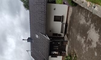 A small house with a gable roof and an open entrance area. The surroundings are green and natural, with a gray sky in the background.