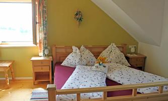 A cozy bedroom with a wooden bed and colorful pillows. The room has a window with a view outside and a small table next to the bed.