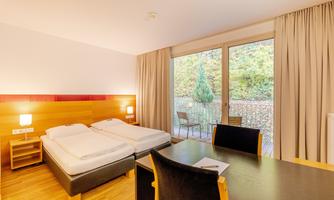 A modern hotel room with two single beds and a desk. Large windows provide light and a view of the outside landscape.
