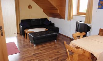 A cozy living space with a black sofa and a wooden table. The walls are light and there are large windows with curtains.