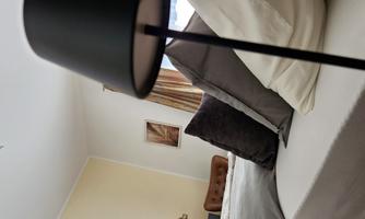 A modern bedroom with a beige tone and elegant furnishings. A nightstand with a black lamp and several cushions on the bed are visible.