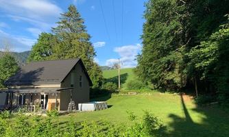 A small house surrounded by lush greenery and trees. In the background, gentle hills and a clear blue sky can be seen.
