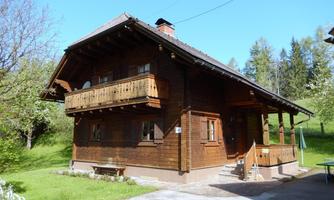 A cozy wooden house with a balcony and a green meadow in the background. The sun is shining and the sky is blue.