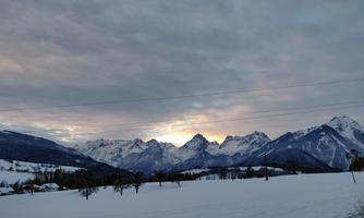 A winter landscape with snow-covered mountains under a cloudy sky. The sun is slowly setting, casting a warm light over the scene.