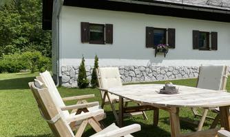 A cozy outdoor area with a wooden table and chairs on green grass. In the background, there is a bright house with window shutters and a flower bench.