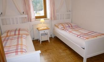A simple bedroom with two twin beds and a small nightstand. The room has a bright, inviting impression with a window and curtains.