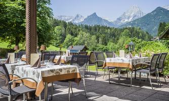 An inviting outdoor area with tables and chairs, surrounded by lush greenery and mountains in the background. The clear sky creates a pleasant atmosphere.