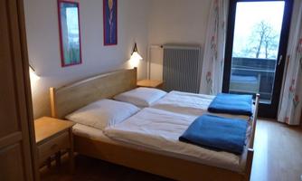 A cozy bedroom with a double bed and two blue blankets. The walls are decorated with colorful pictures, and there is a balcony with a view.