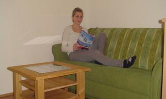 A woman is sitting comfortably on a green sofa, reading a magazine. In front of her stands a small wooden table.