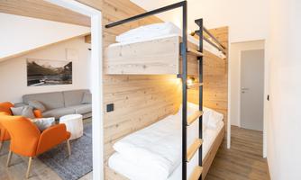 A modern bedroom with a wooden bunk bed and a cozy living area. The walls are light, and there is a comfortable sofa as well as orange chairs.