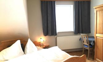 A bright room with a double bed and a chair at a table. Large windows let in a lot of daylight.