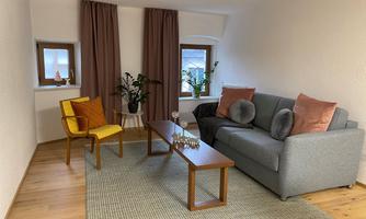 A cozy living room with a gray sofa and a yellow chair. There are plants and a wooden table on a carpet.