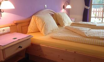 A cozy bedroom with a double bed and checked pillows. The walls are painted in a soft purple, and there is a window with curtains.