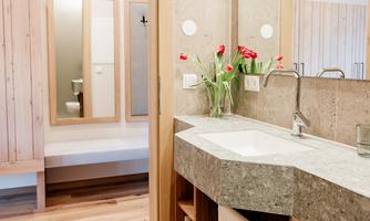 A modern designed bathroom with a stone sink and a wooden shelf. Fresh flowers are placed on the sink and a large mirror is visible in the background.