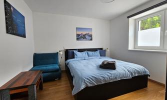A modern bedroom with a large bed and blue bed sheets. Pictures are hanging on the wall and the room has a cozy armchair and a small table.