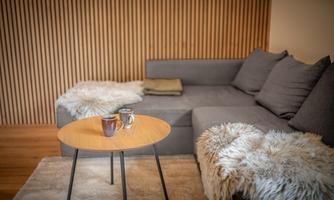 A cozy living room with a gray sofa and soft blankets. On a round table, there are two cups and a decorative fur mat.