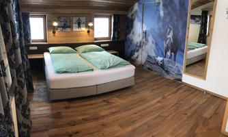 A cozy bedroom with a double bed and wooden flooring. On the wall hangs a picture of a horse, and there are large windows that let in natural light.
