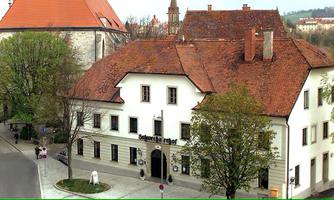 A historical building with a red tiled roof and white walls. It is surrounded by trees and people can be seen on the street.