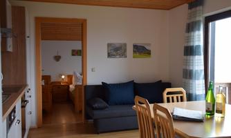 A cozy living area with a sofa and a dining table. In the background, you can see a bedroom and windows with a view outside.