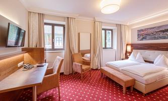 A cozy hotel room with two beds and a desk. The walls are light, and there are large windows with curtains.