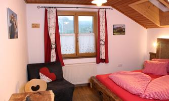 A cozy bedroom with a large bed and red pillows. There is a comfortable armchair and a window with curtains.