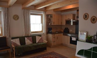 A cozy living room with a kitchenette and a couch. Wooden beams and large windows create a bright atmosphere.