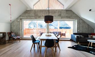 A modern living room with high ceilings and large windows. The dining table in the center is surrounded by stylish chairs and offers a view of the terrace.