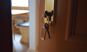 A key is hanging in a door lock. In the background, a room with furniture and natural light is visible.