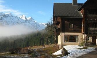 A rustic wooden house in the mountains with a snow-covered ground. In the background, impressive mountain peaks and a clear sky can be seen.