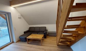 A modern living room with a comfortable sofa and a wooden table. A wooden staircase leads to the upper floor, while large windows provide plenty of light.