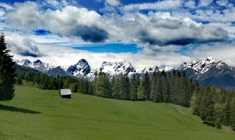 A green meadow with a small wooden house. In the background, snow-covered mountains rise under a cloudy sky.
