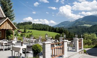 A sunny terrace with tables and chairs, surrounded by green meadows and mountains. The sky is blue with some white clouds.