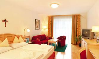 A cozy hotel room with a double bed, a red seating area, and a large window. The decor is bright and friendly with wood and warm colors.