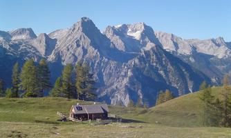 A cozy cabin stands on a green meadow with majestic mountains in the background. The sky is clear and nature appears untouched.