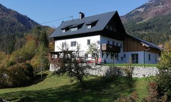 A modern house in the mountains, surrounded by trees and nature. The facade is light with a dark roof and a balcony.