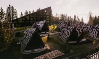A modern hotel building surrounded by stylish, triangular cabins in nature. Tall trees and a calm sky create a relaxed atmosphere.