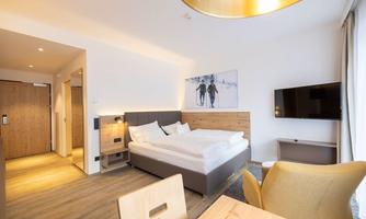 A modern hotel room with a double bed, a television, and a seating area. The walls are light, and the decor is simple and cozy.