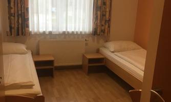 A simple room with two beds and a chair. The windows are covered with curtains and let in a lot of light.