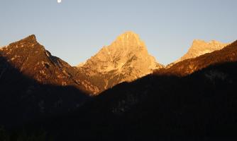 An impressive mountain landscape with snow-covered peaks in the evening light. The moon is visible in the sky.