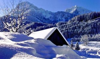A snowy landscape with a small wooden house and snow-covered mountains in the background. The sky is clear and bright blue.