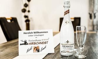 A welcome sign stands on a table, next to a bottle of sparkling wine and two glasses. The sign welcomes guests to the holiday home HIRSCHNEST.