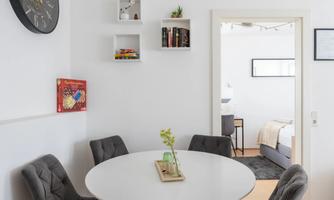 A modern dining area with a round white table and four gray chairs. On the wall, there is a clock and shelves with decorations.