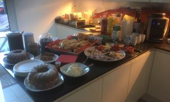 An inviting kitchen with various treats on a table, including cakes, breads, and snacks. There are also drinks and dishes available.